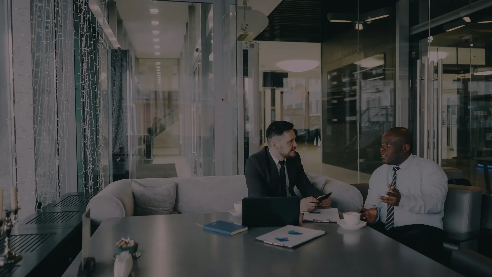 Two business professionals discussing work at a table in a modern office lounge, with a laptop, documents, and coffee cups.