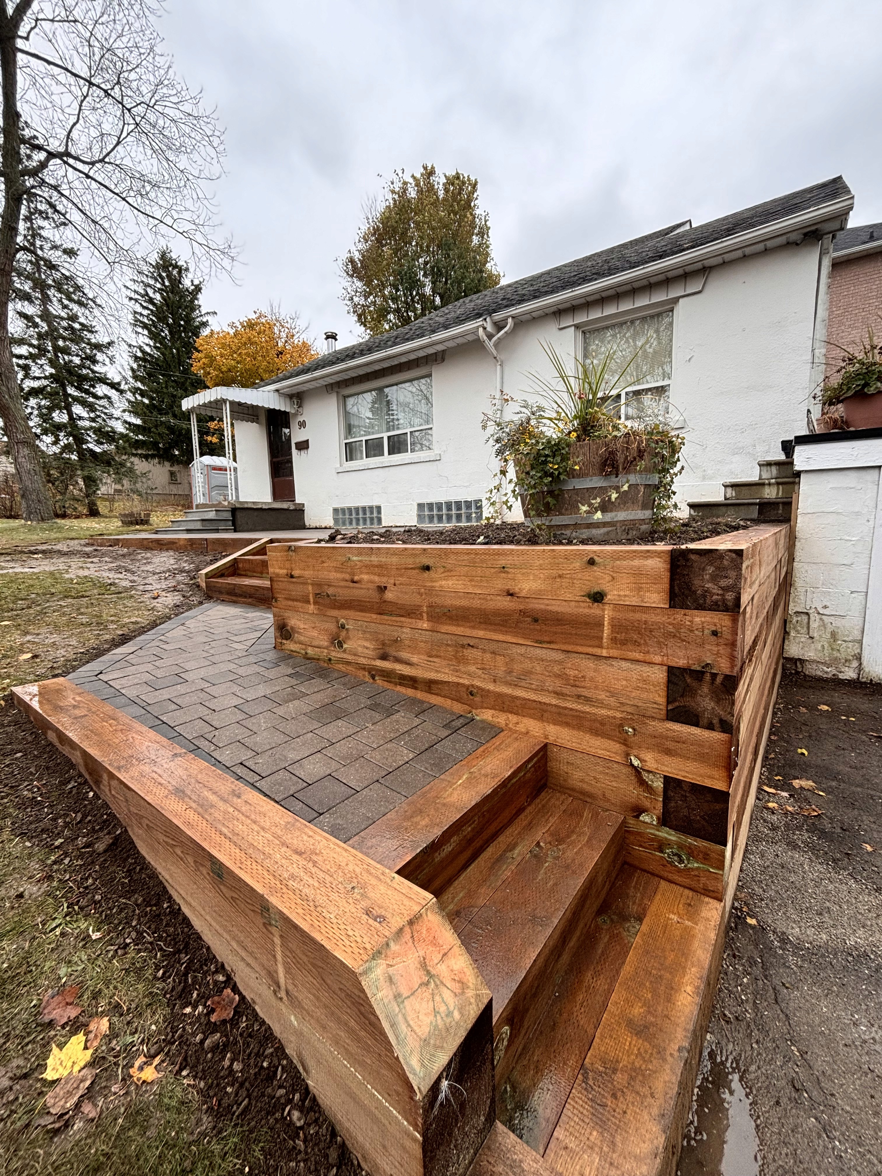 A close-up view of wooden steps leading to a house, with autumn foliage in the background under cloudy skies.