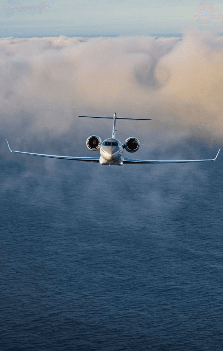 Aerial view of a private jet flying over the ocean, with clouds visible. Plane viewed head-on.