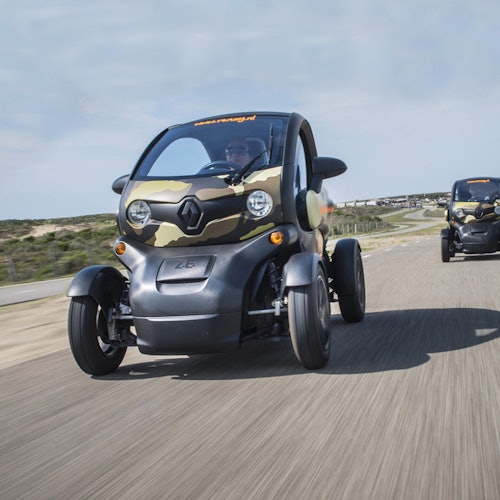 Dua kendaraan listrik kecil dengan kamuflase melaju di jalan dengan latar belakang pemandangan.