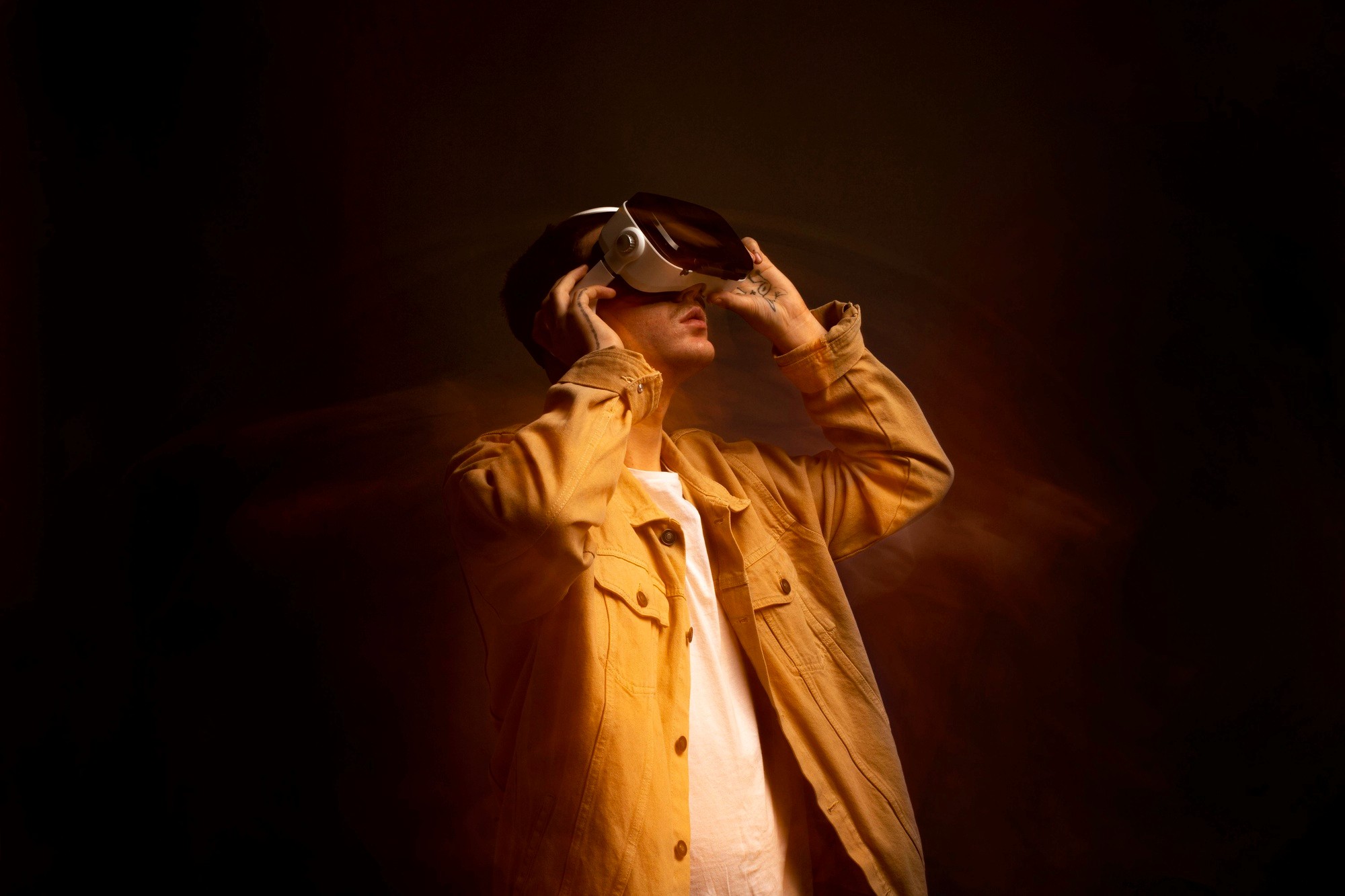 A young man wearing a modern white VR headset, looking upwards in a cinematic studio setting with warm orange lighting and a smoky background.