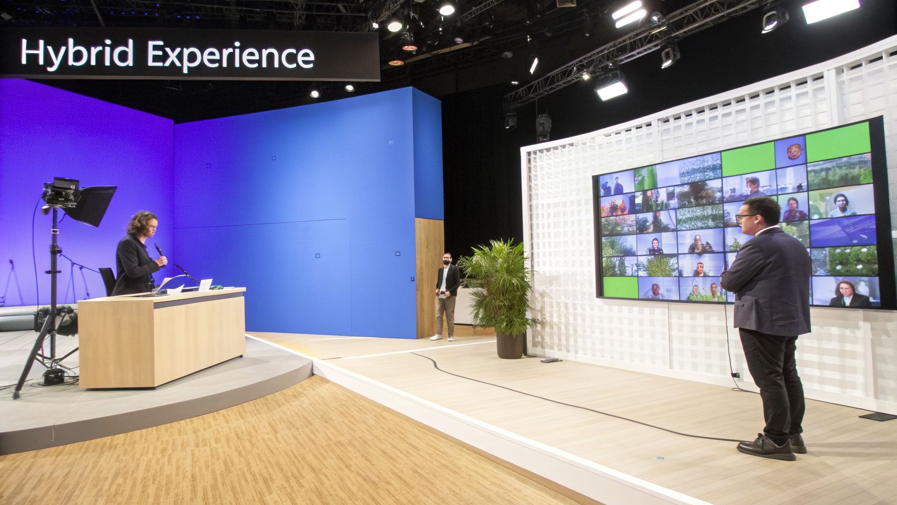 A modern hybrid event space featuring a speaker, stage, and large screen with virtual participants.