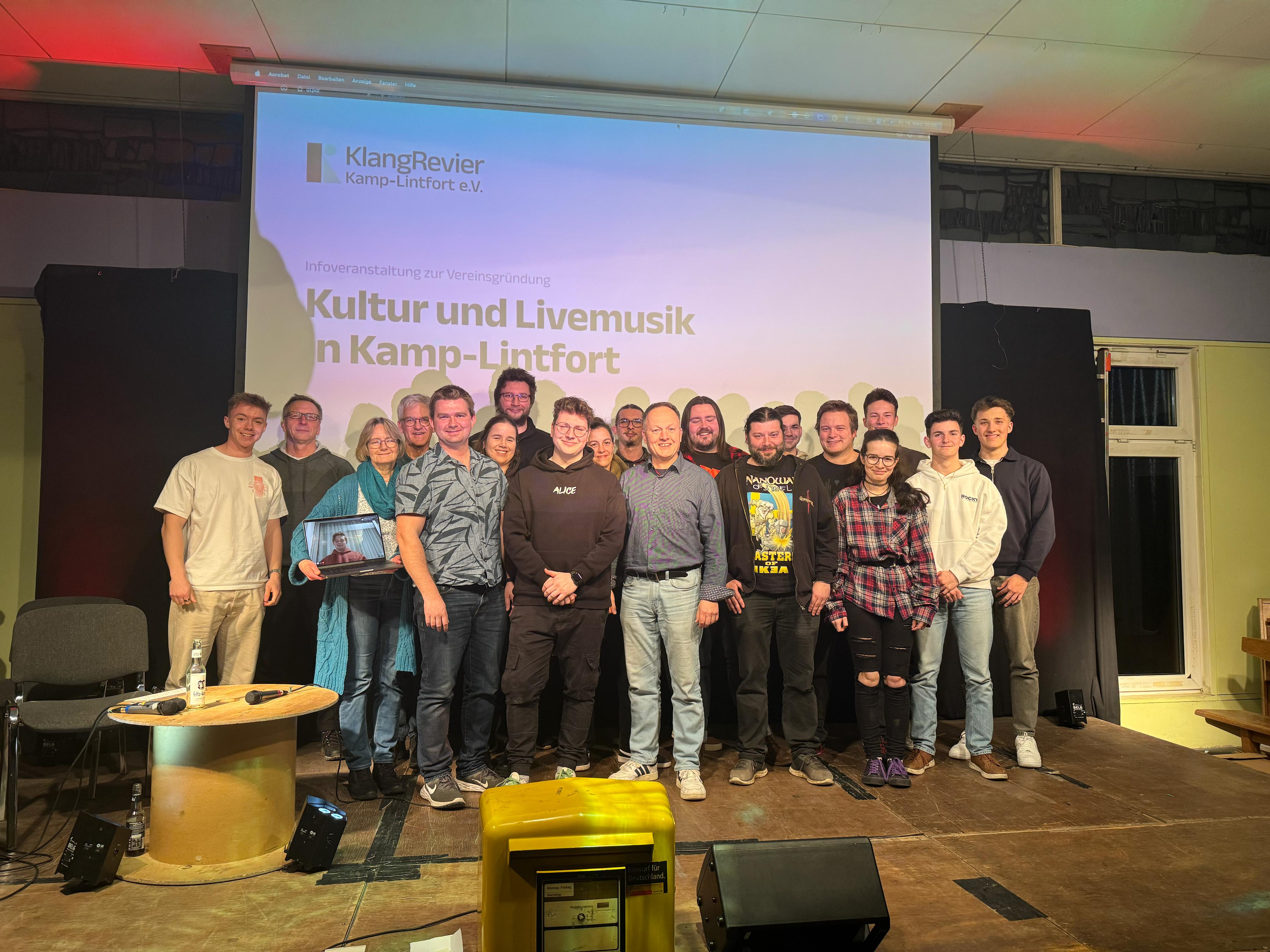 Mehrere Personen des KlangRevier-Teams stehen gemeinsam auf einer Bühne vor einer großen Leinwand mit der Aufschrift ‚Kultur und Livemusik in Kamp-Lintfort‘. Die Gruppe wirkt freundlich und präsentiert sich als Teil des Festivals.