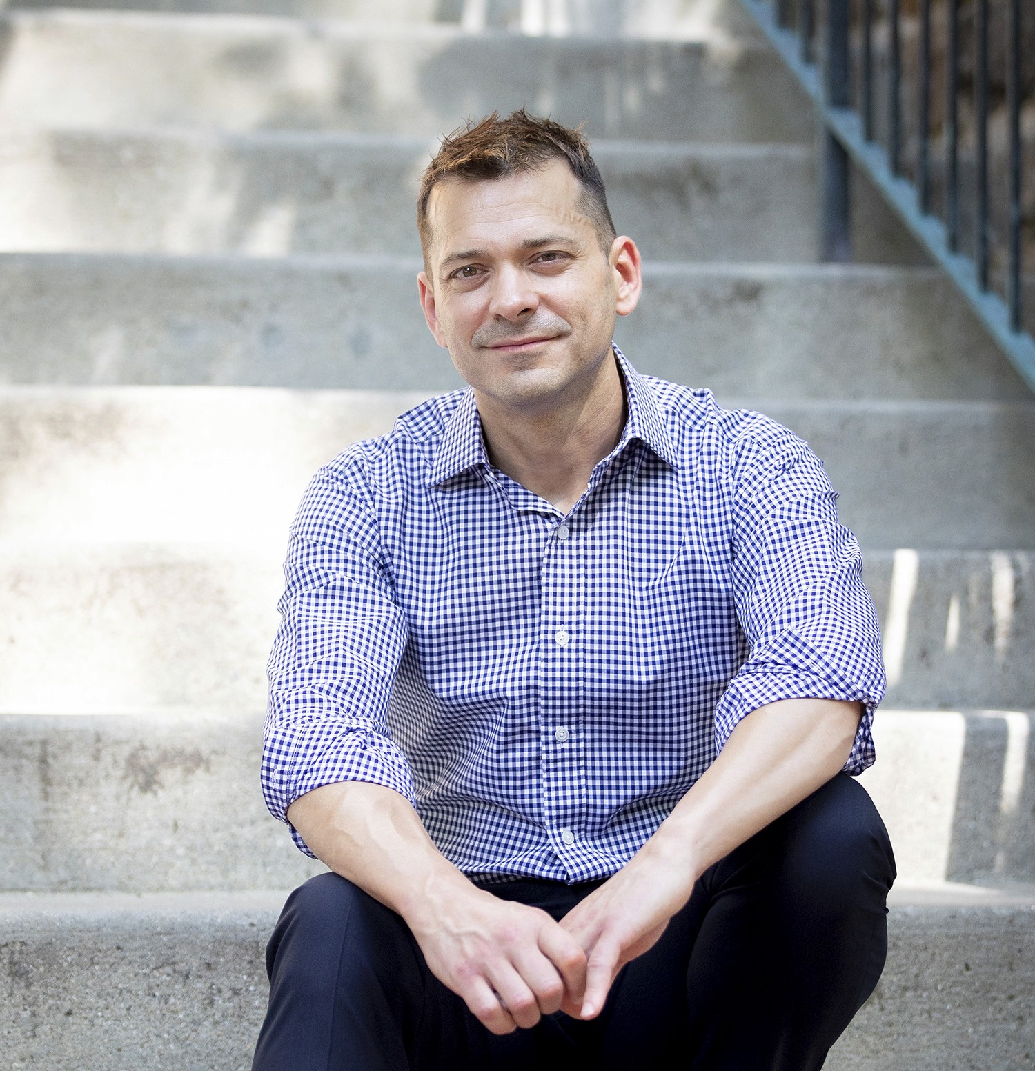 A man sits casually on stairs, smiling while wearing a light blue shirt and black pants.