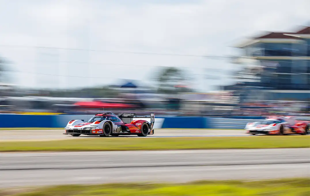 Two red, black, and white race cars compete on the Sebring track, one chasing the other, with a motion-blurred background emphasizing the intense speed.