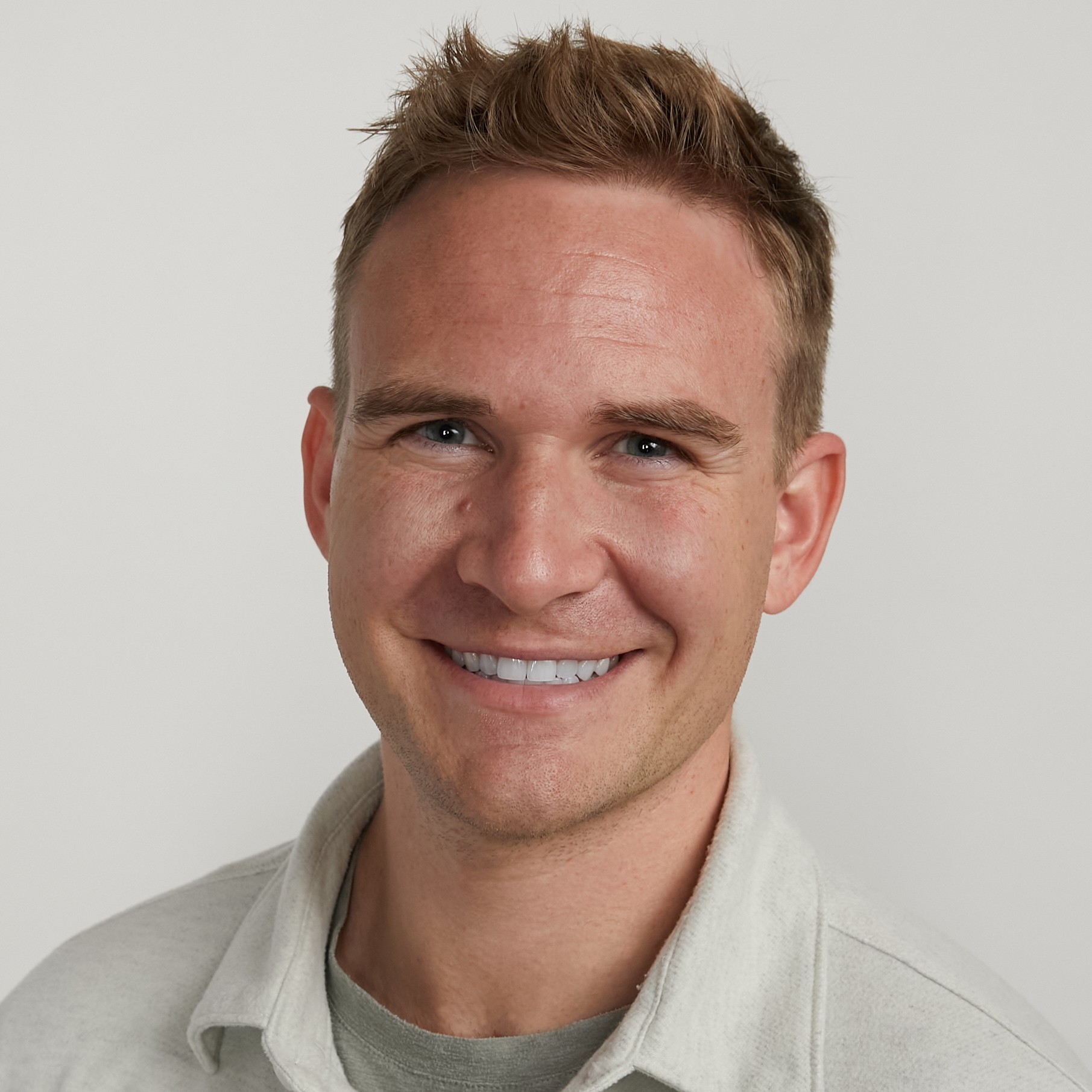 A smiling person with curly hair sits at a desk, looking at a laptop against a dark background.