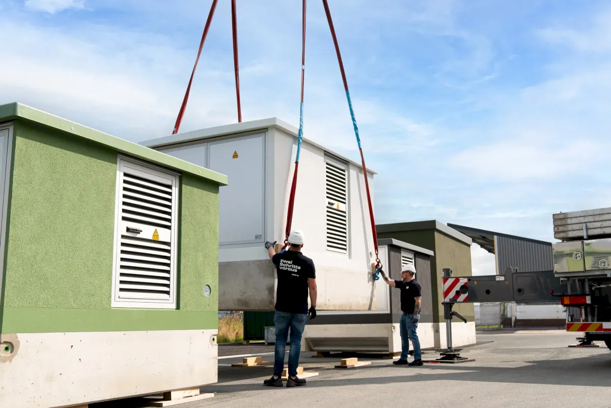 Zwei Arbeiter heben mit einem Kran einen großen weißen Container an.