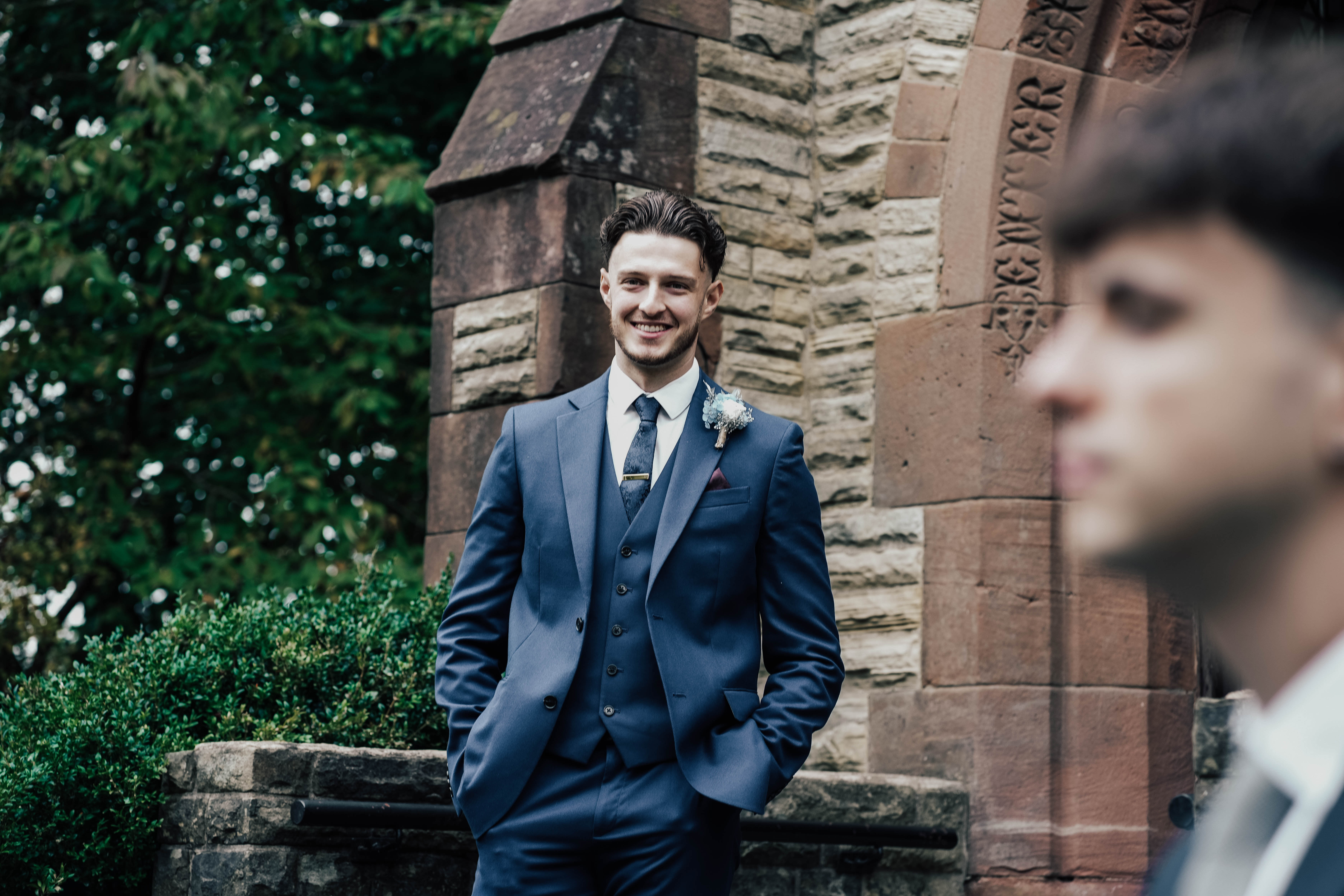 A young groomsman in a blue three-piece suit smiles while standing with his hands in his pockets outside a stone church. Another guest is visible in the out-of-focus foreground.
