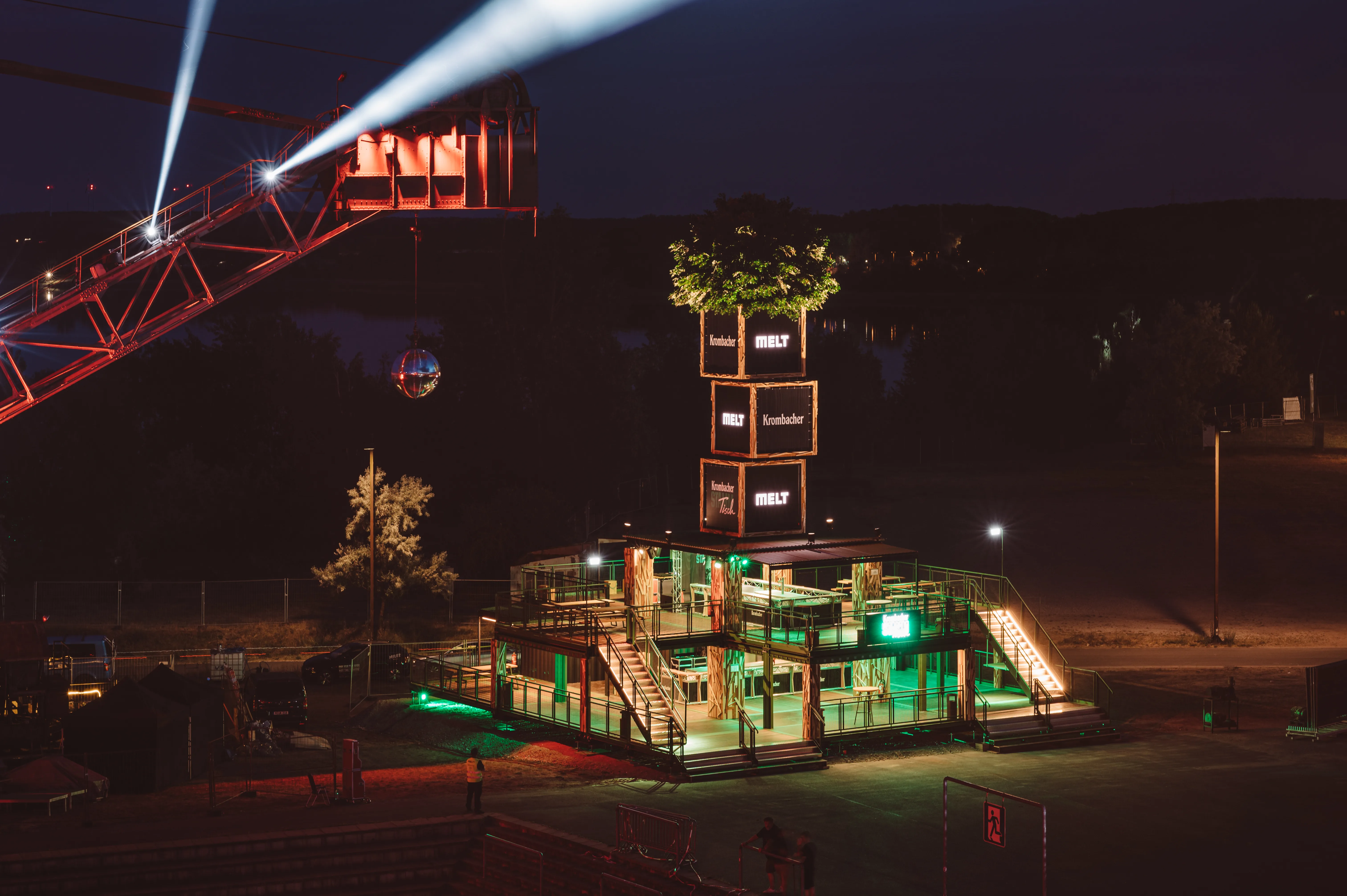 KROMBACHER festival stand at night with green-lit two-storey scaffold structure, illuminated tower of branded crates topped by a tree and light beams from a red crane.