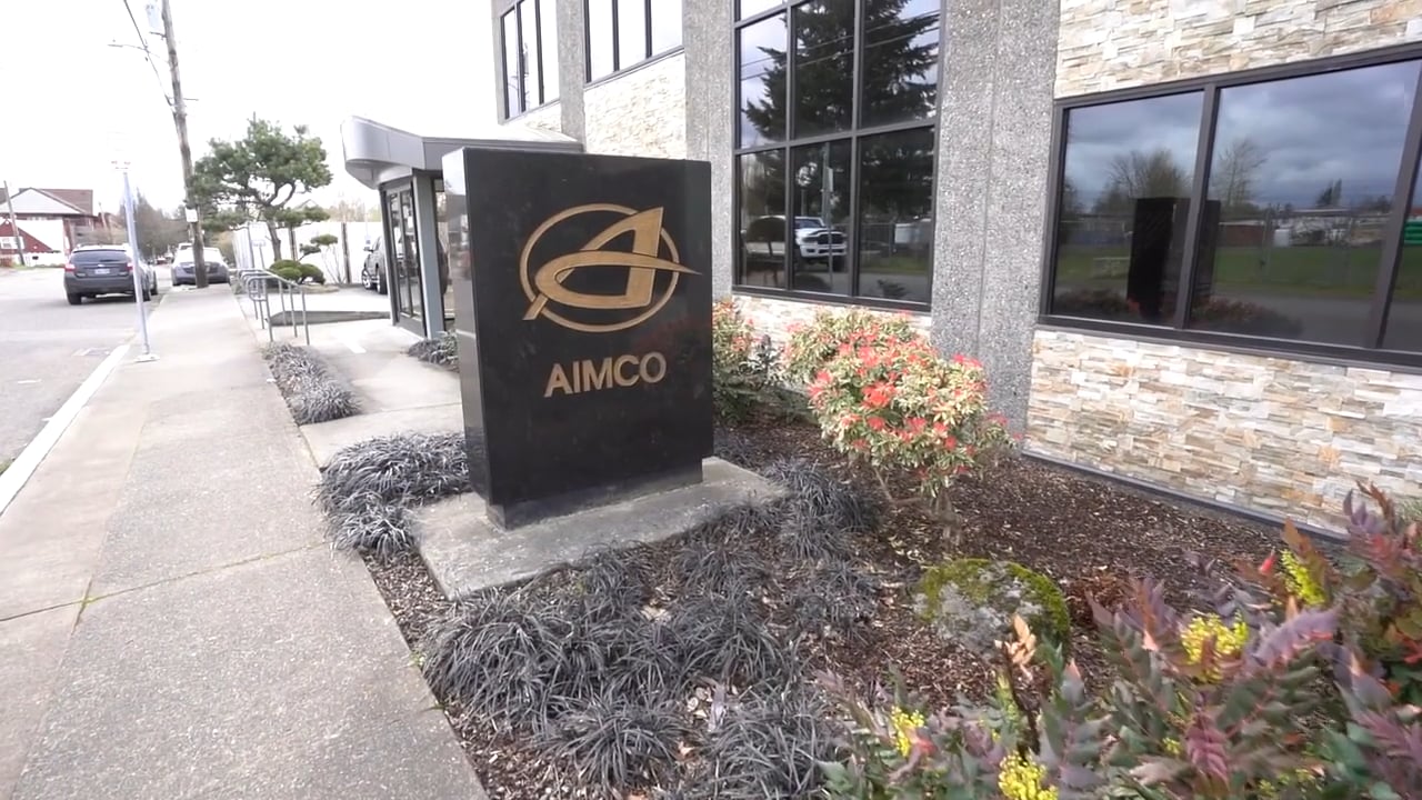 Image of a building sign with a logo, situated near a sidewalk and landscaped area. Windows visible in background.