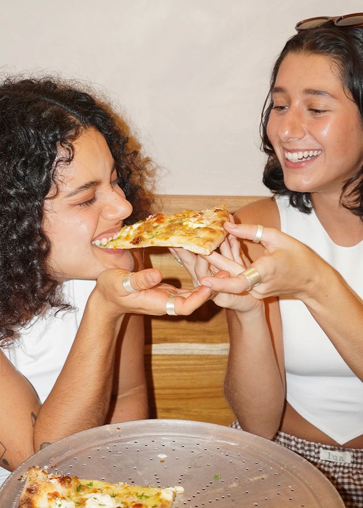 Two women sharing a slice of pizza indoors wearing silver rings