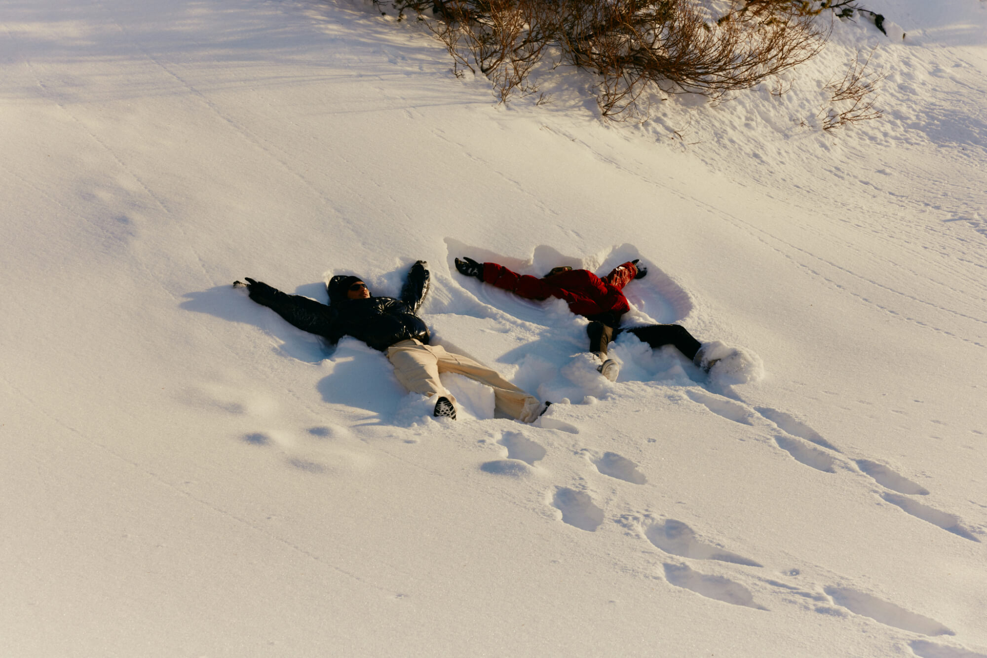 Two people lie in the snow making snow angels, one in a dark jacket, the other in red. Footprints lead to them. The scene feels joyful and playful.