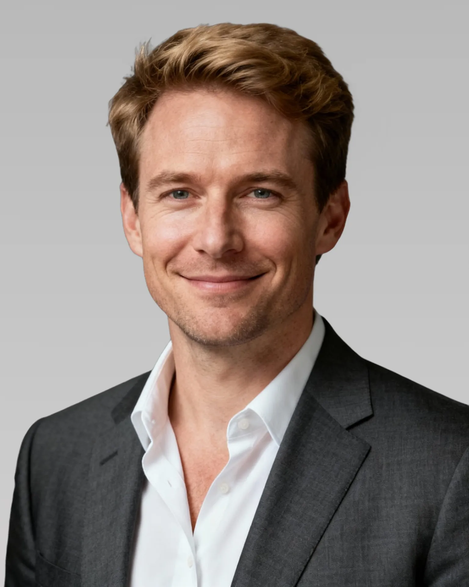 A man with blonde hair and a gray suit smiles warmly against a neutral background.