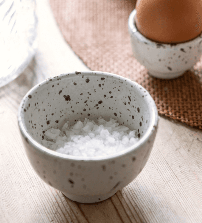 Petit bol en céramique mouchetée gris et brun contenant du sel de mer, avec coquetier et œuf en arrière-plan sur table en bois clair