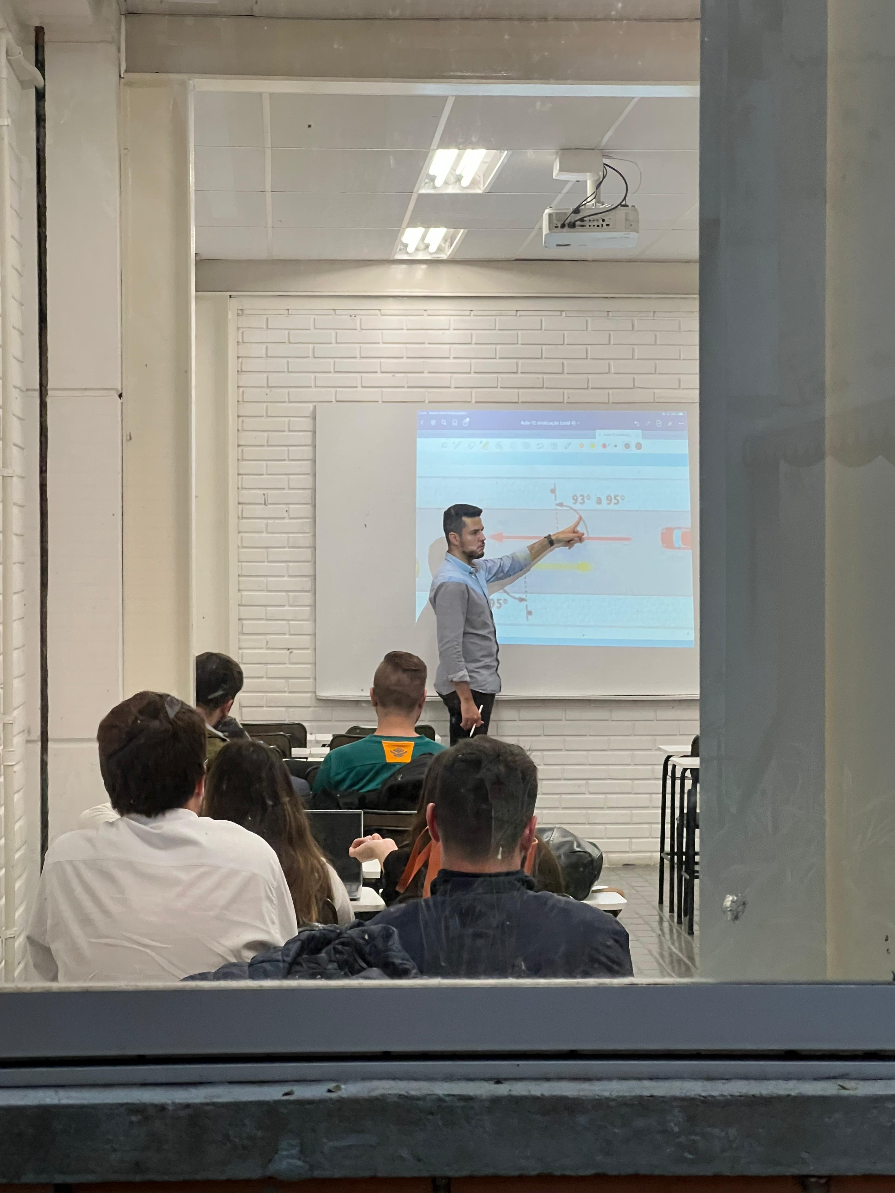 Professor Eduardo Alexandre Kruger ministrando aula teórica de engenharia civil, apontando para uma projeção de traçado geométrico de rodovia.