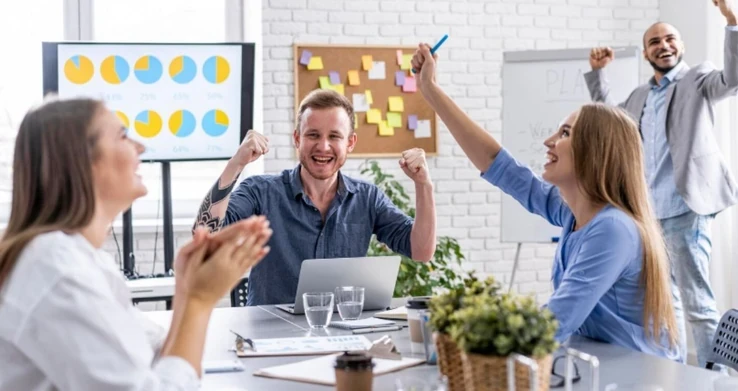 Equipe comemorando de braços erguidos em volta de uma mesa, com um quadro de avisos ao fundo