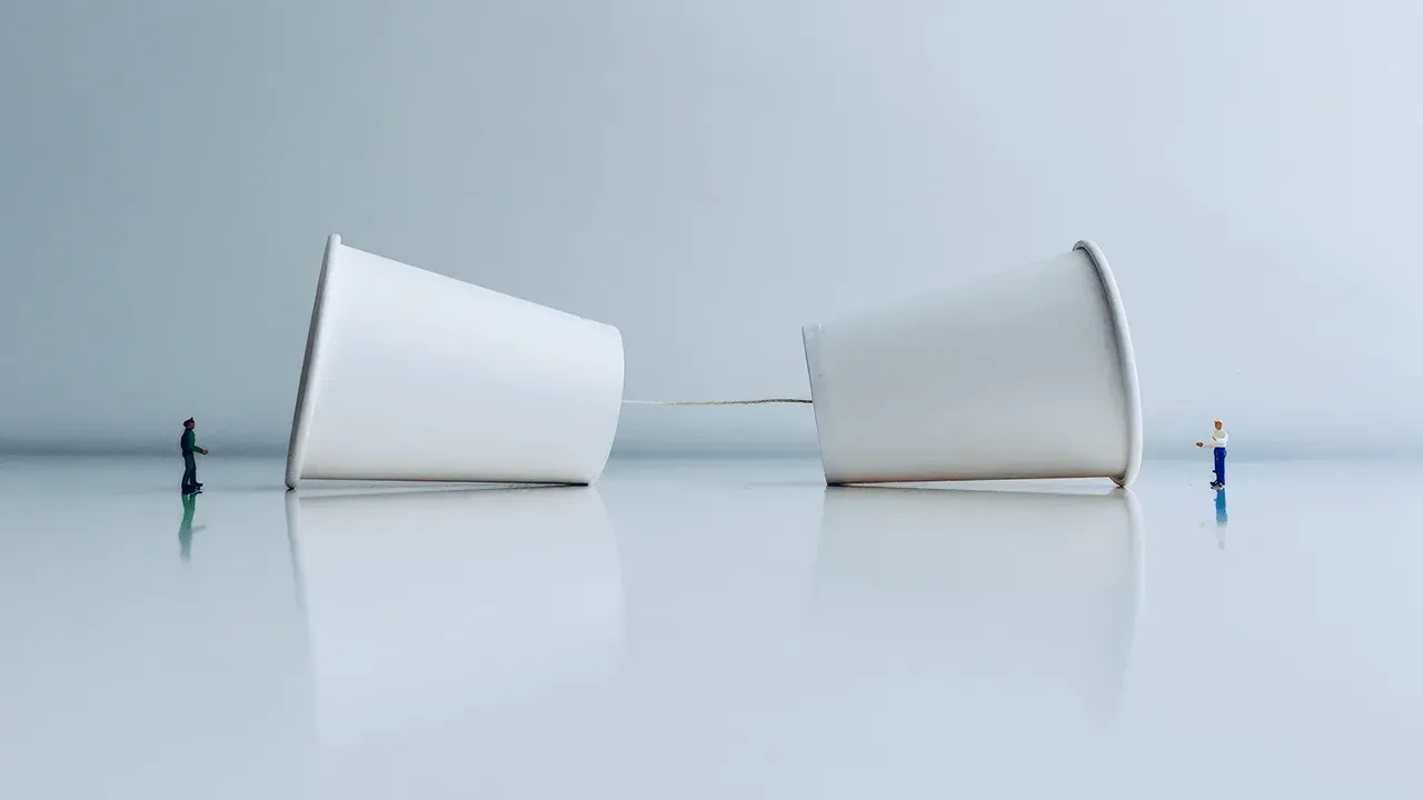 Two paper cups connected by a string, with miniature figures near each cup, symbolizing communication, connection, or a breakdown in dialogue.