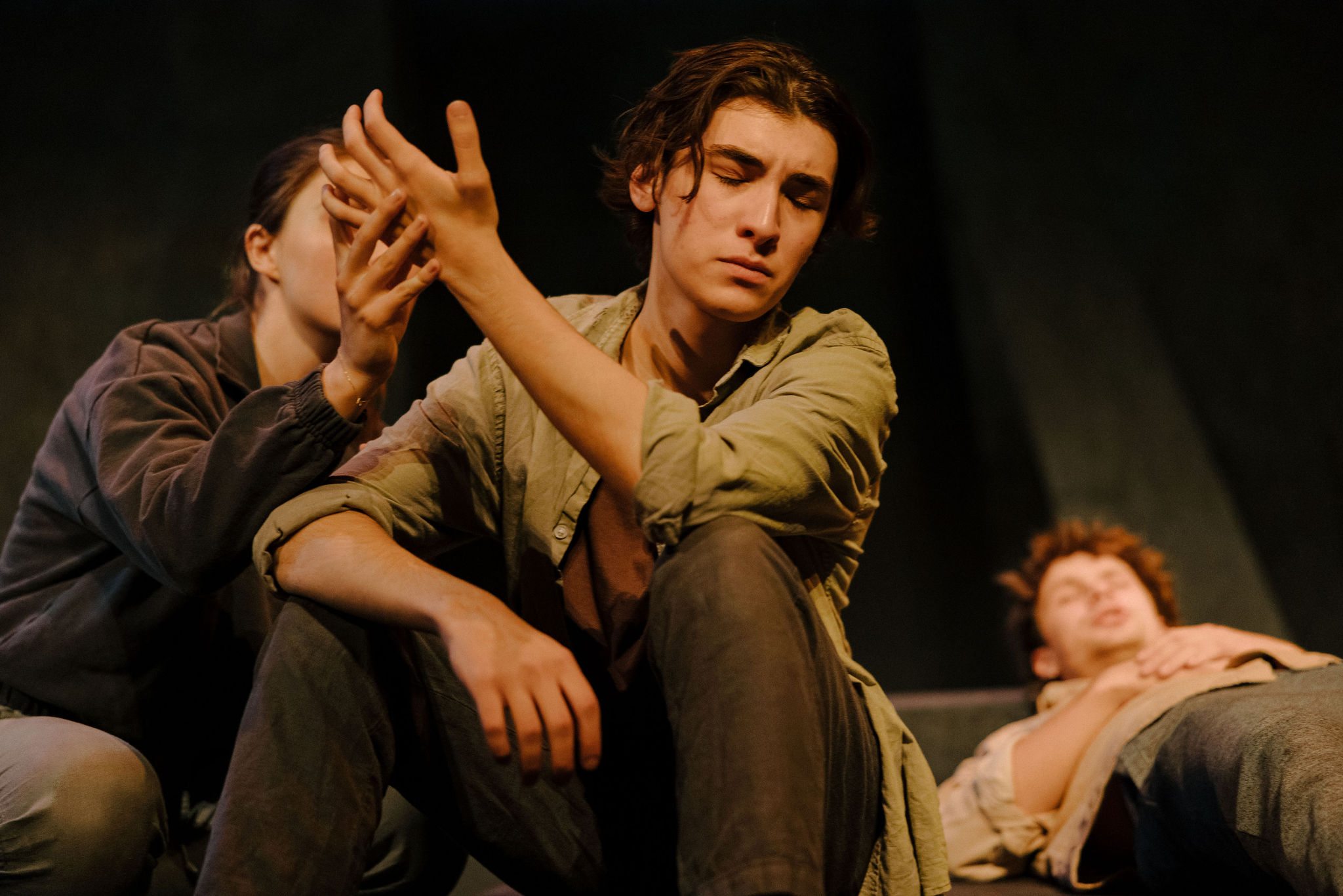 A performer sits with eyes closed while another gently lifts their hand, with a cast mate resting behind them on the floor.