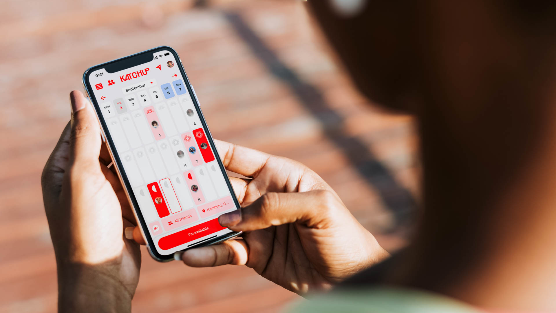 A person holds a smartphone displaying the KATCHUP app. The screen shows September 2nd through September 7th, with options for 'FRI', 'SATURDAY', 'MON', 'TUES', 'WEDNESDAY', 'THURSDAY', 'FRIDAY', 'SATURDAY', 'SUNDAY', 'MONDAY', 'TUESDAY', 'WEDNESDAY', 'THURSDAY', 'FRIDAY', 'SATURDAY', 'SUNDAY', and 'MONDAY'. The text 'I'm available' appears below the date. The background is blurred, showing a wooden surface.