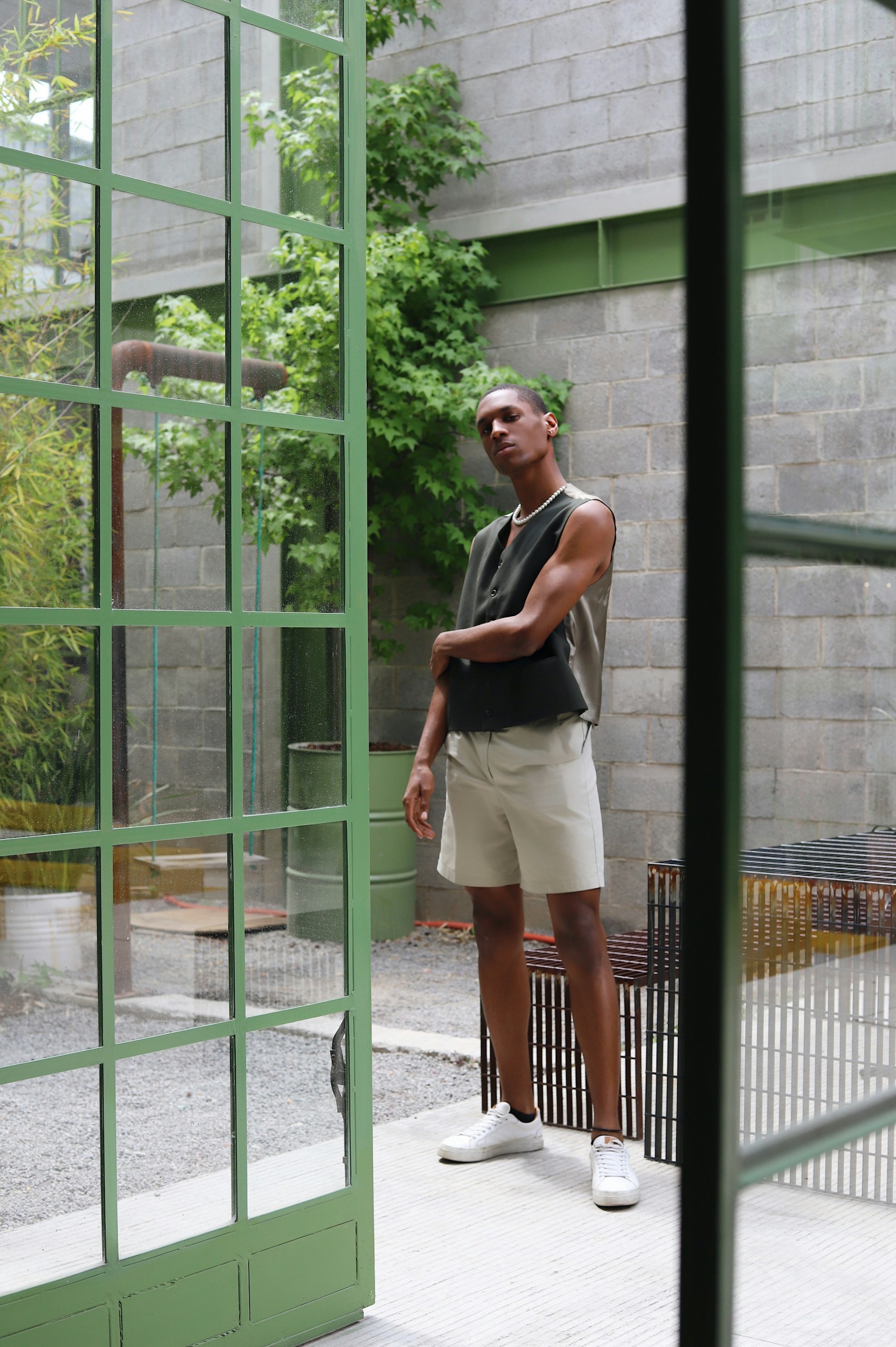 A full-length shot of a man standing in an outdoor courtyard seen through a large, green-framed glass door. He is wearing a dark vest, light-colored shorts, and white sneakers against a background of concrete blocks and green foliage.