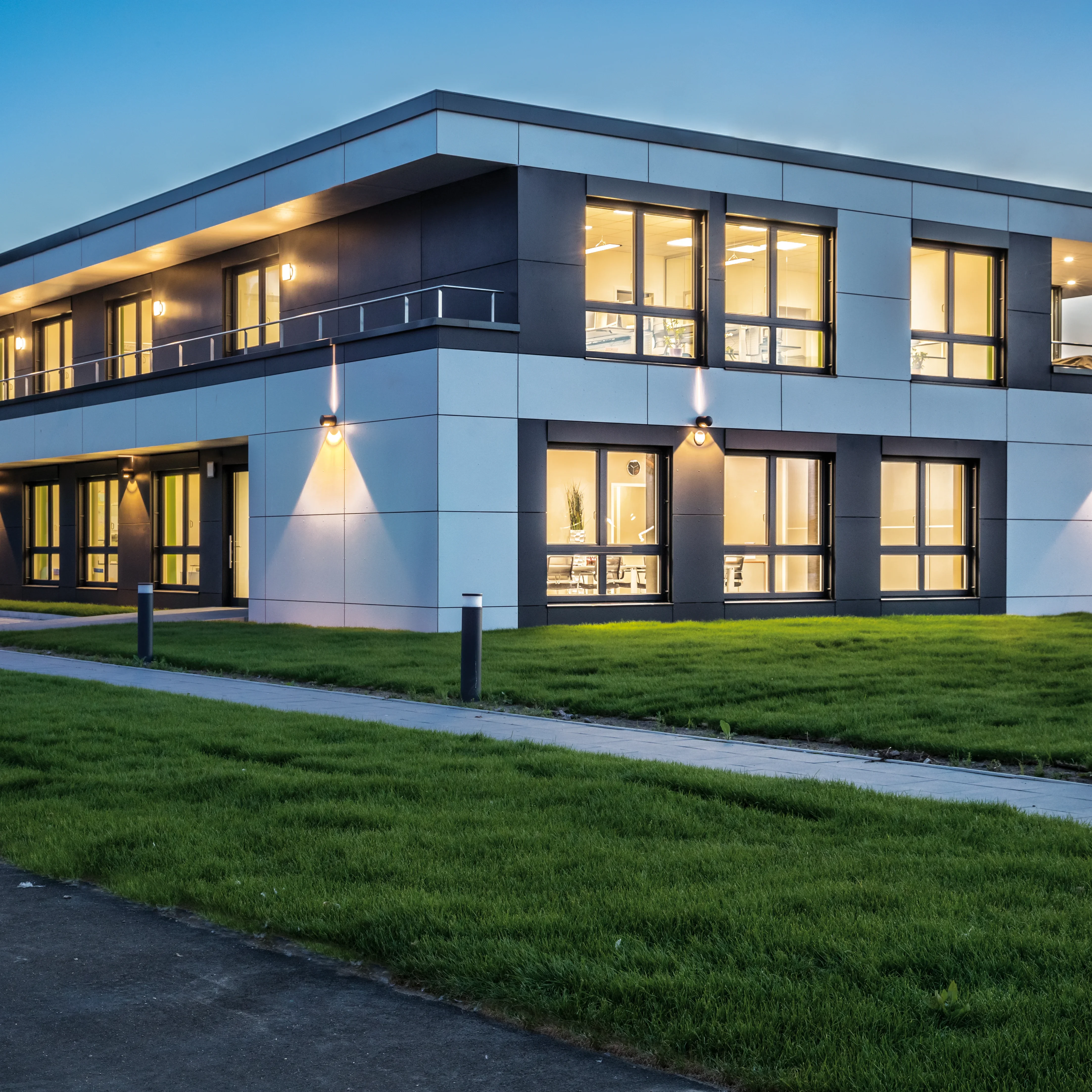 Moderne zweistöckige Bürogebäude mit hell beleuchteten Fenstern und dunkler Fassade, umgeben von sattgrünem Rasen.