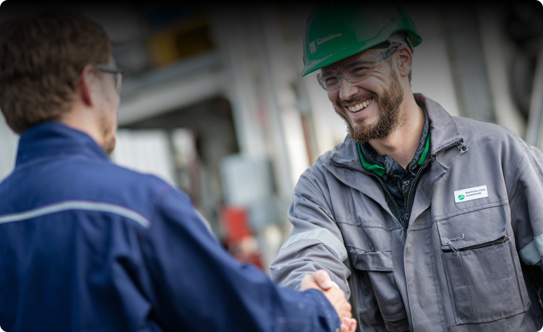 A smiling man with a beard, wearing a green hard hat, safety glasses, and a grey work jacket, shakes hands with another person in a blue uniform in an industrial setting.
