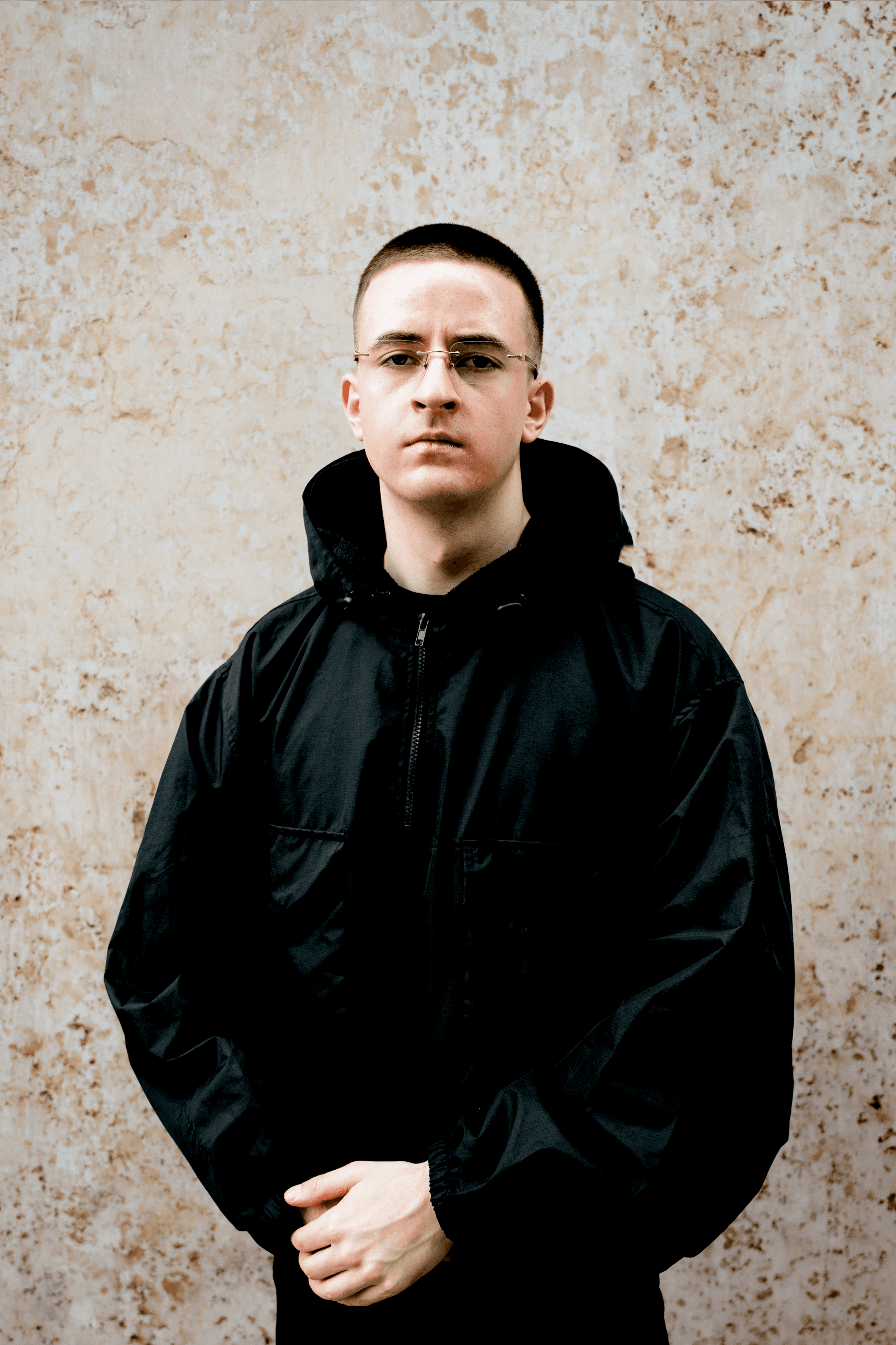 Elyas, a German-based music producer, wearing a black jacket against a textured beige wall, gazing confidently at the camera.