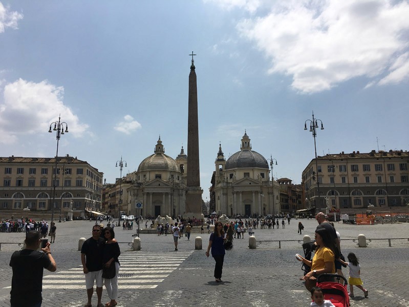 A photo showing a paved square market with an obelisk like structure and some old buildings. Also there are people walking by and taking pictures.