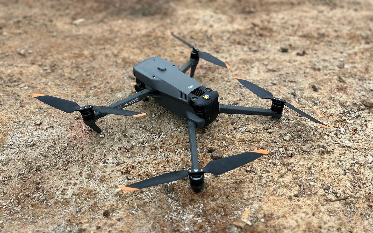 Close-up de um drone profissional cinza da marca DJI com hélices de pontas alaranjadas, repousando sobre o solo de terra batida de uma obra. A imagem destaca o equipamento utilizado para a Captura da Realidade aérea.