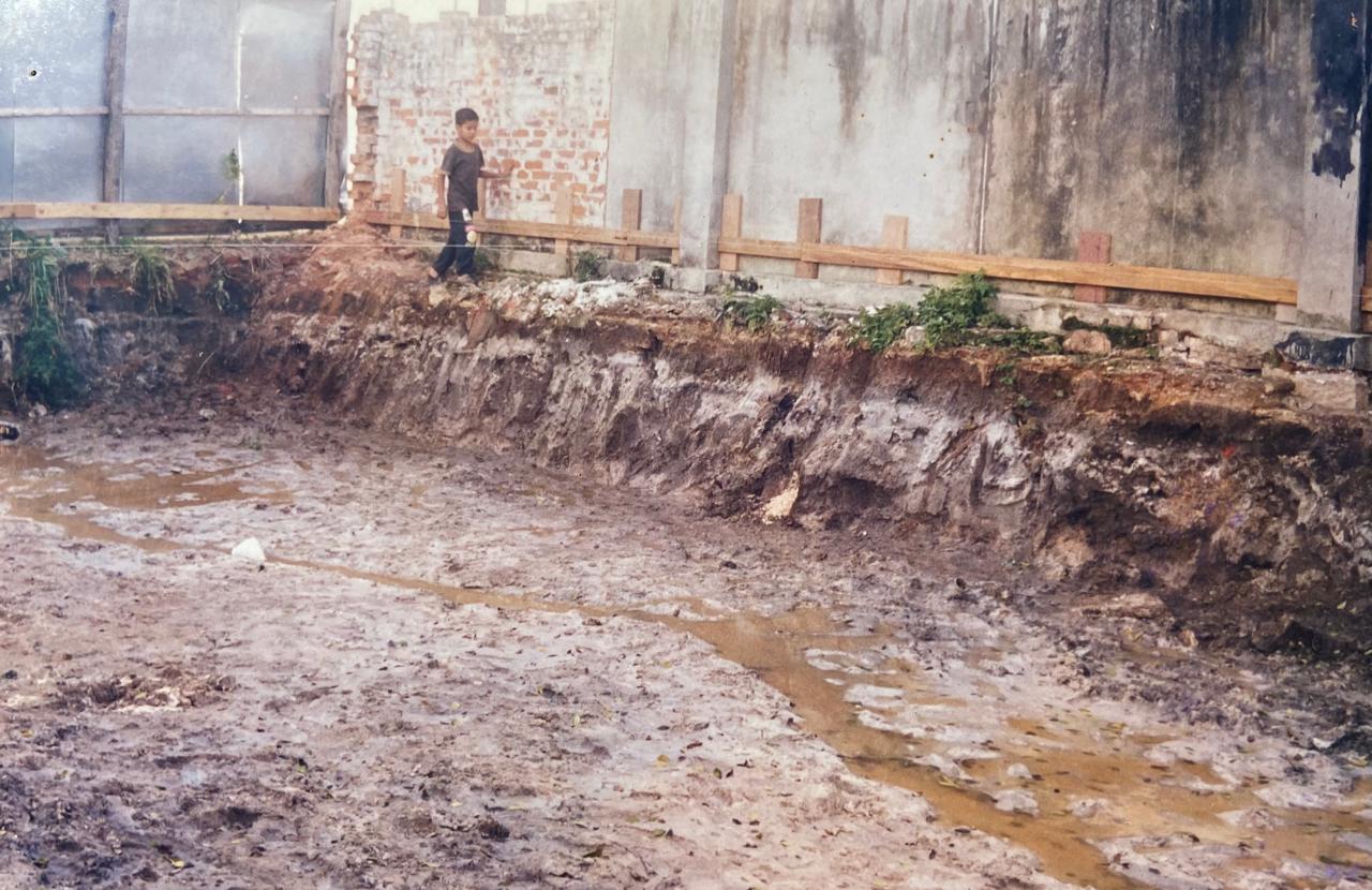 Escavação de solo e preparação de terreno em canteiro de obras para fundação de engenharia civil. Ao fundo, parede de tijolos e marcações de madeira.