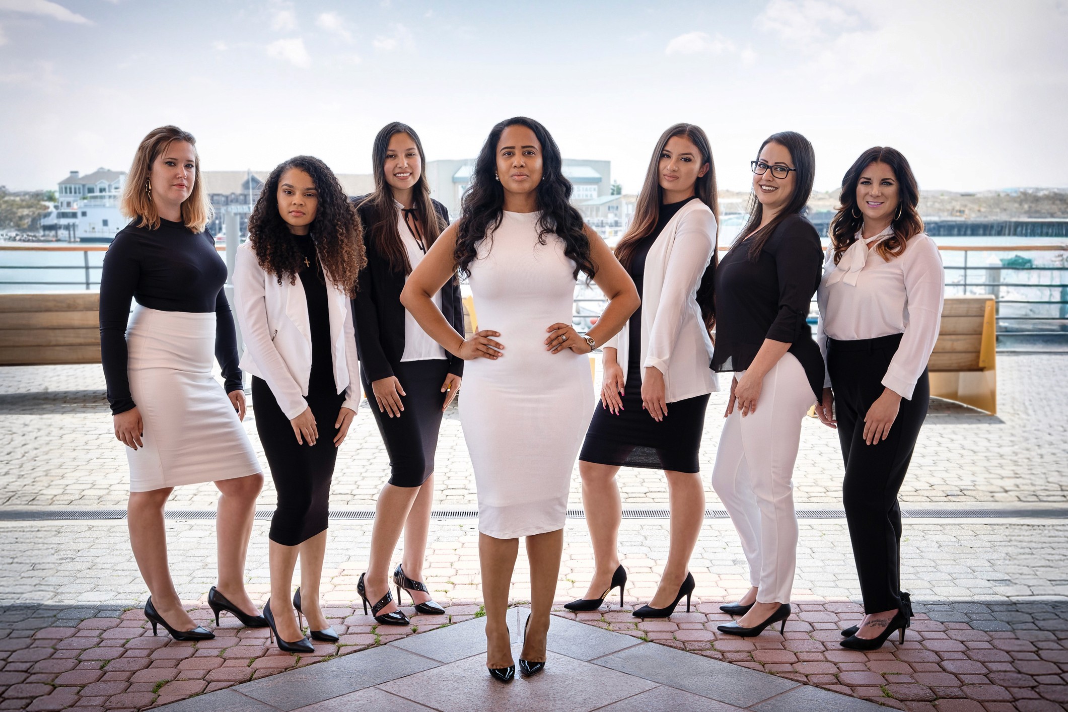 Team branding photo of seven professional women in black and white outfits standing confidently on waterfront patio with modern marina in background