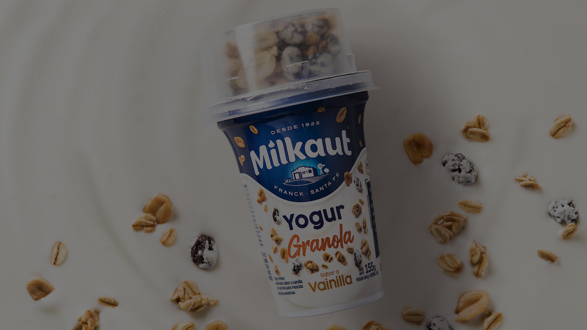 Plastic cup of Milkaut vanilla yogurt topped with granola. Scattered oats and granola pieces surround the cup on a white surface.