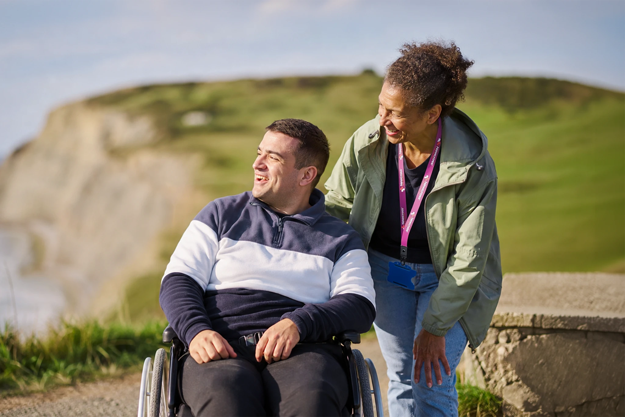 Support worker walking alongside a smiling wheelchair user on a coastal path, both enjoying the view together.