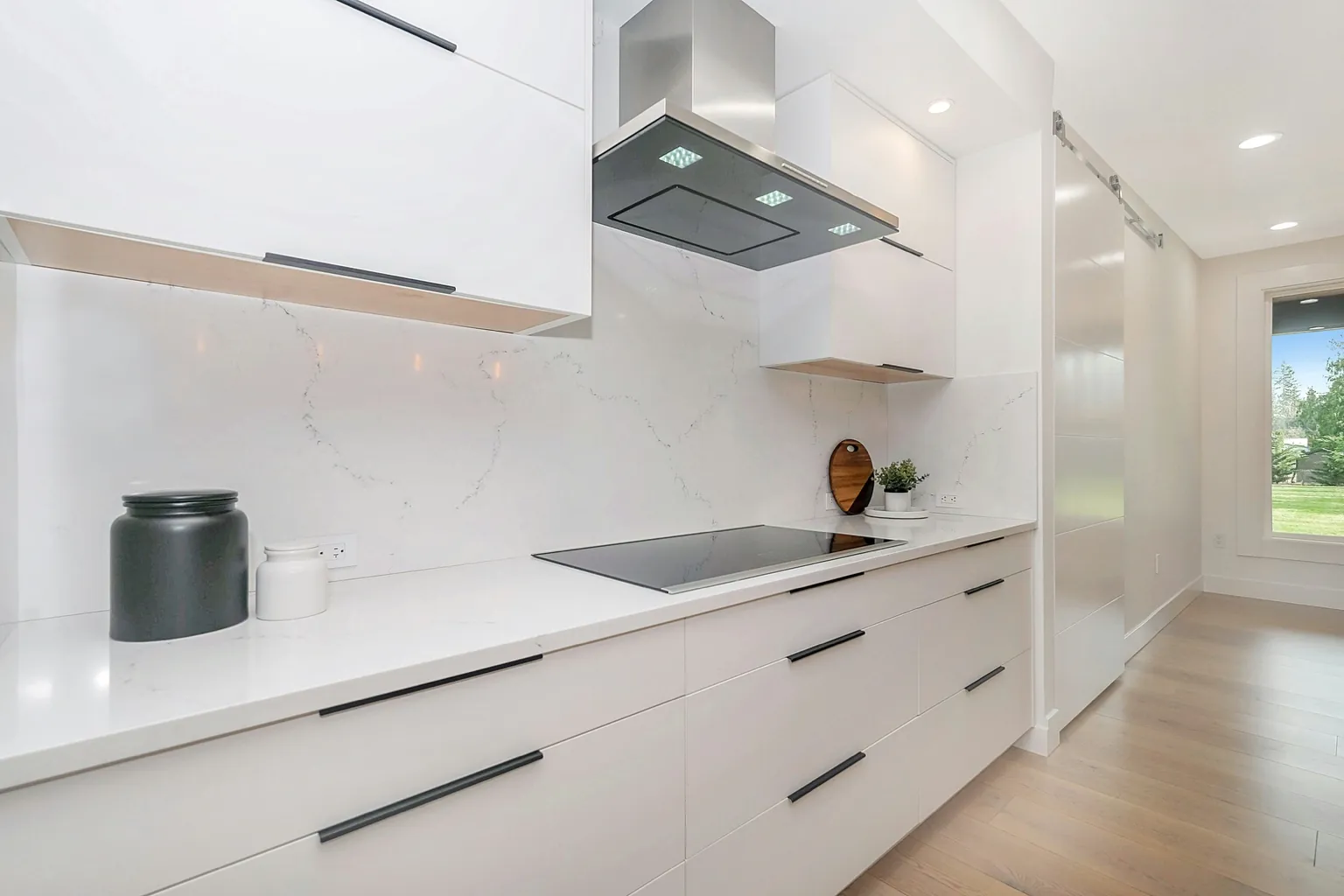 Cucina moderna contemporanea con mobili bianchi lucidi, piano di lavoro chiaro, cappa in acciaio inox e illuminazione a soffitto.