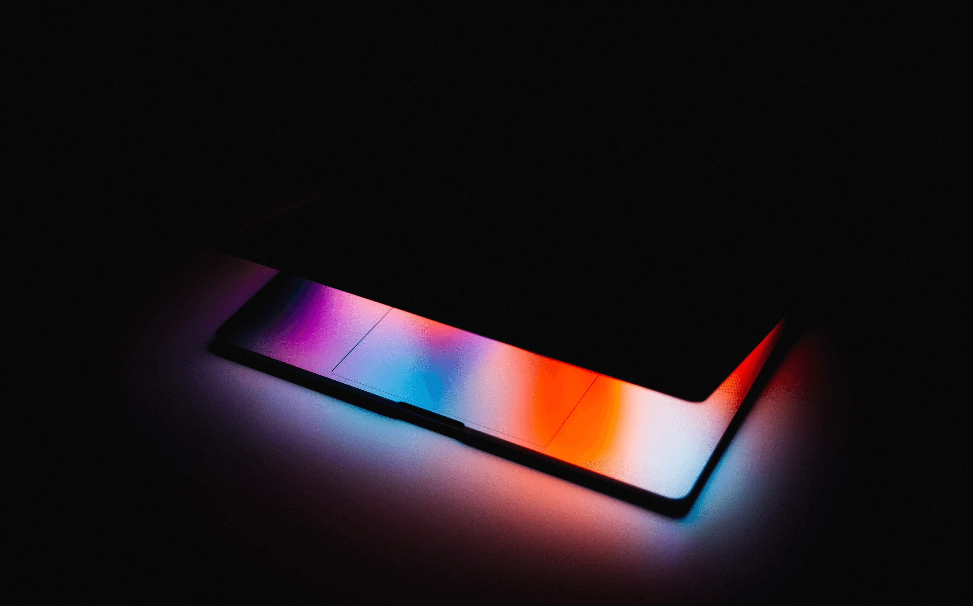 Laptop about to open, only the colourful light of the screen is visible.