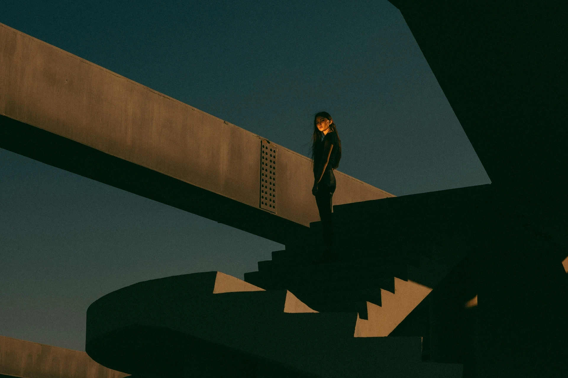 Person steht auf einer Treppe zwischen klaren, geometrischen Betonstrukturen im Abendlicht, reduzierte Architektur mit starken Linien und Kontrasten.