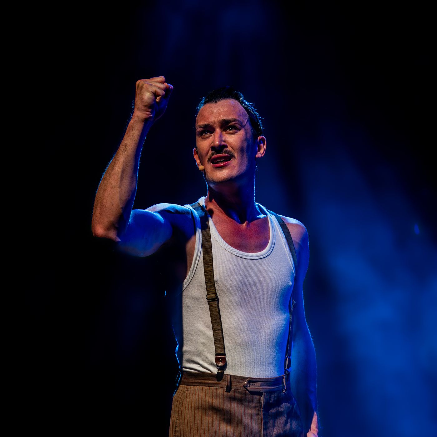 James Warner, en débardeur et bretelles, le poing levé, chante dans une lumière bleue