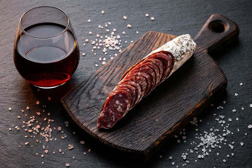 Sliced black truffle salami on a wooden board with a glass of red wine, from Porcellino Artisanal Meats.