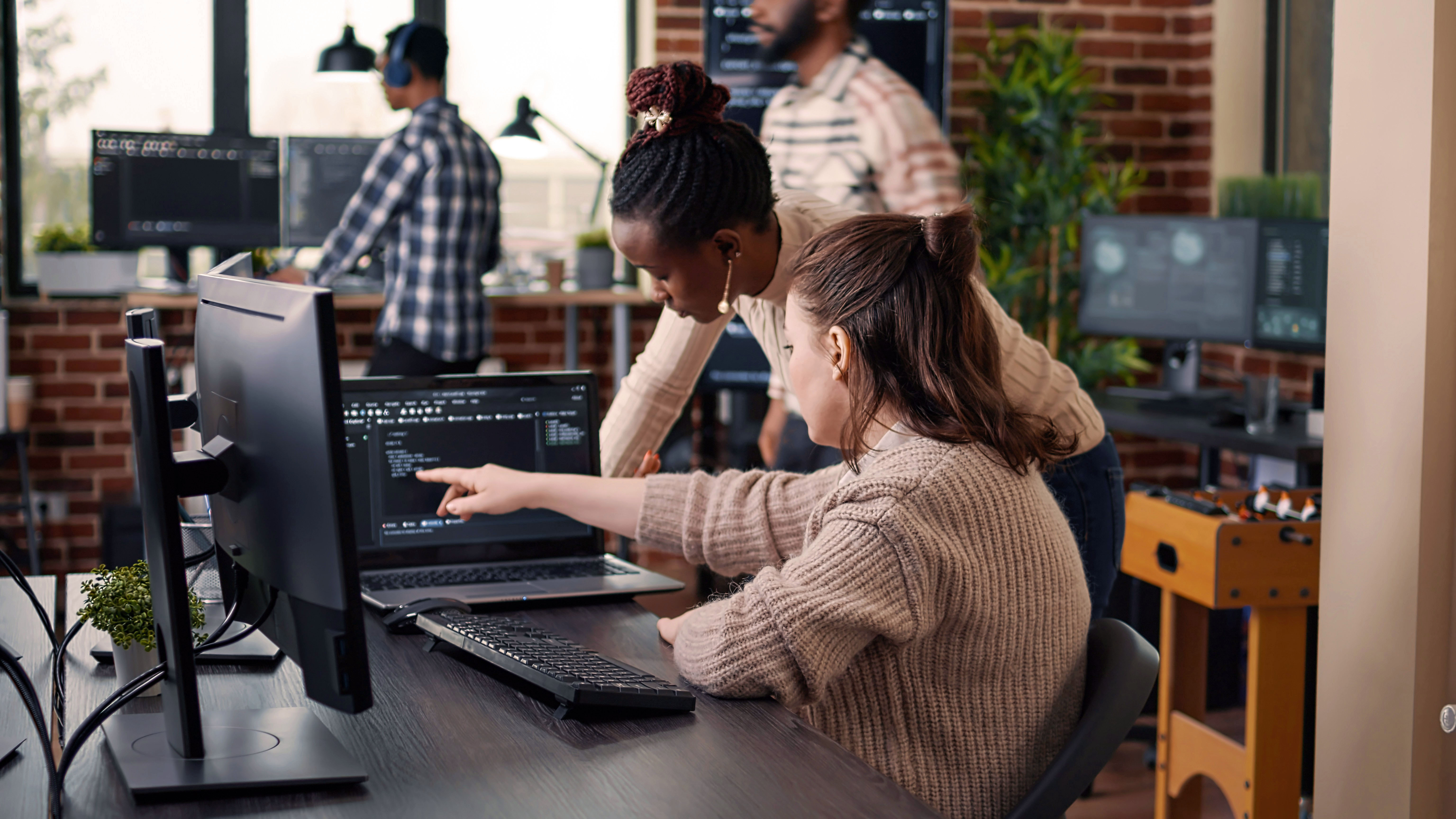 Two people working at computers in a bright office space, with one person assisting the other.