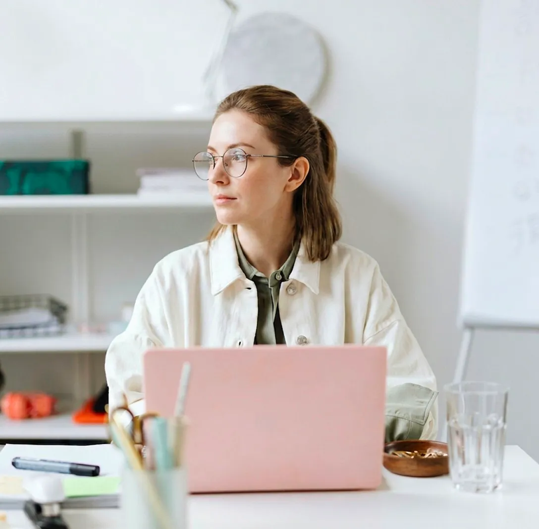A woman with glasses at a desk with a laptop, looking up thoughtfully