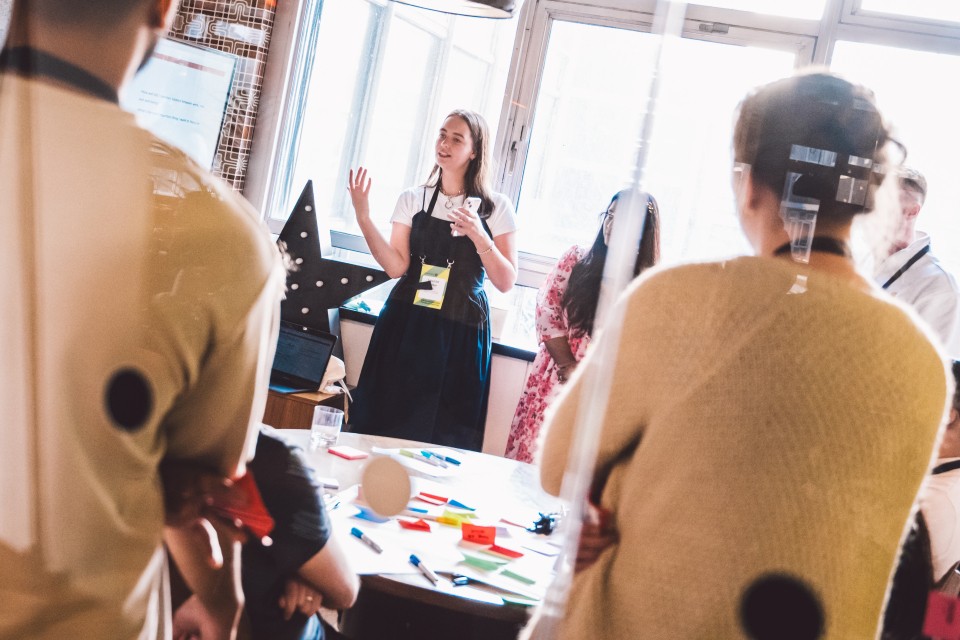 A group of people stand around a table covered with colourful sticky notes and workshop materials, while one person at the front speaks to the room. The scene appears to take place in a bright space with large windows, and the group is gathered for a collaborative session or presentation.