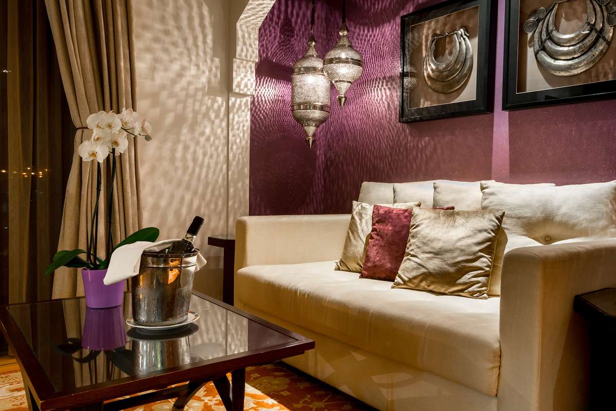 A wine bottle and glass sit on a dark coffee table in front of a cream sofa against a textured purple wall with art.