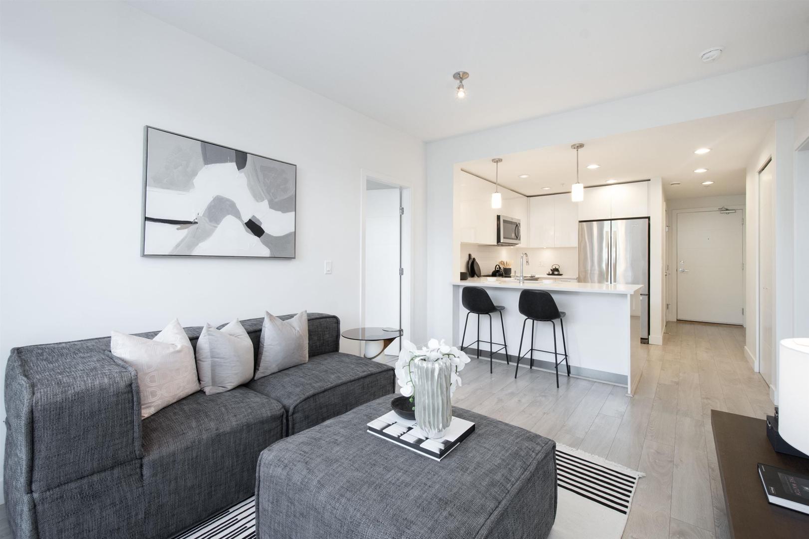 A modern living room with a gray couch, sleek decor, and an open kitchen in the background. Bright and airy atmosphere.