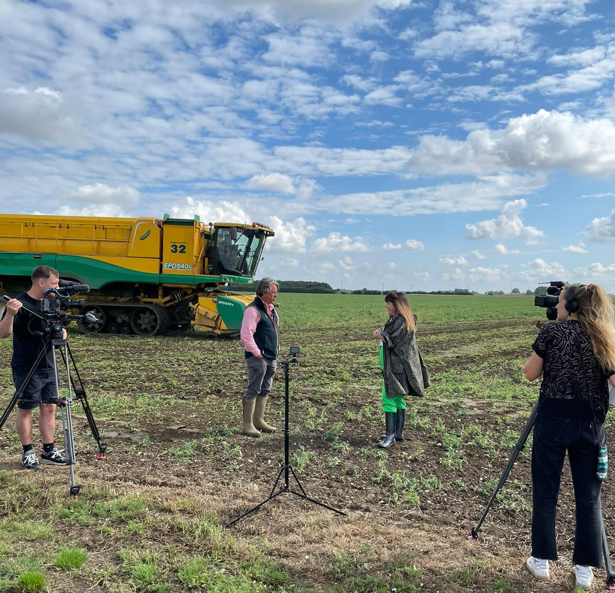 Briony Williams interviewing Stephen Francis. They are in a field with a yellow and green Ploeger harvester in the background. The sky is blue with dappled cloud
