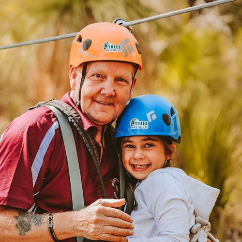 在一个户外探险乐园里，一位成人和一名儿童都佩戴着头盔和安全带，微笑着站在一起。