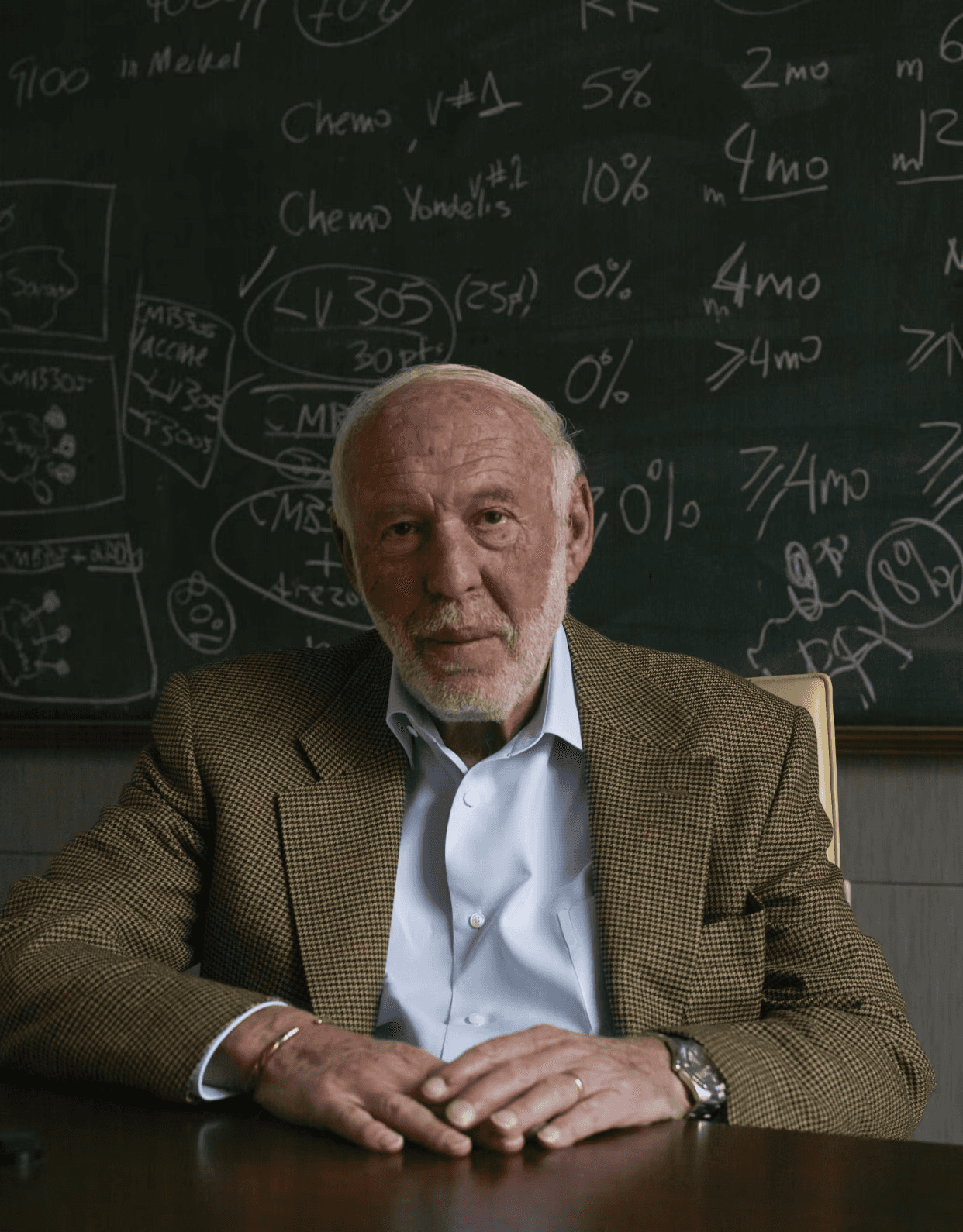 Nobel laureate Dr. David Baltimore seated in front of a chalkboard with medical formulas and vaccine notes.
