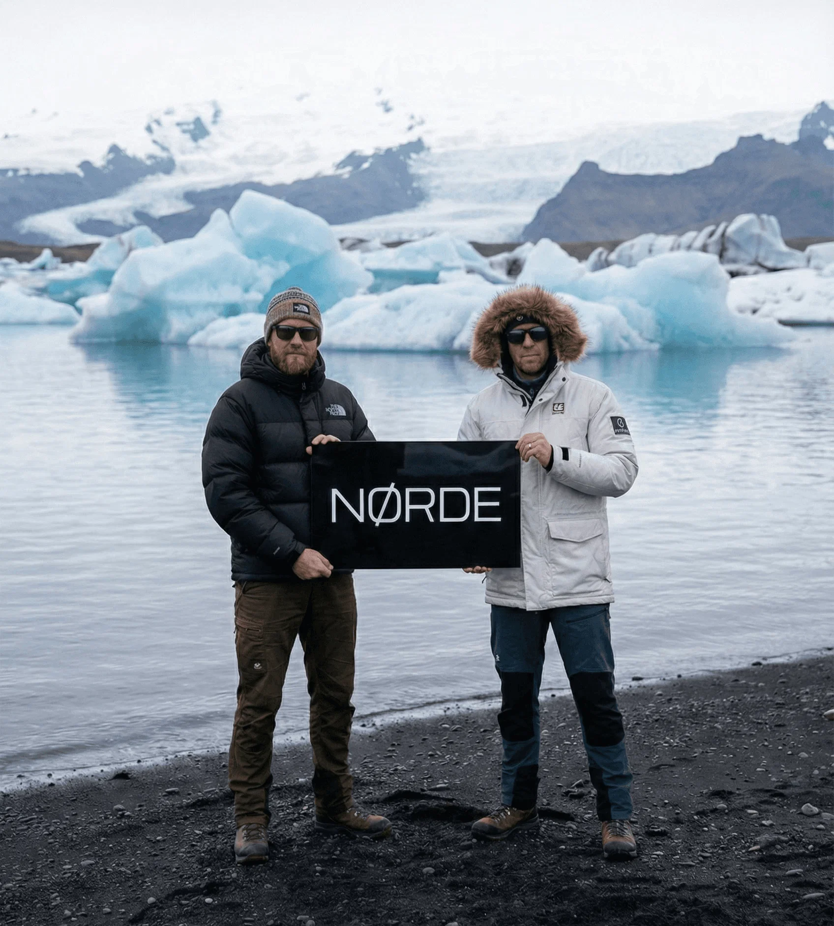 Two men in winter jackets standing on a black sand beach holding a black sign, with blue icebergs and a glacier in the background.
