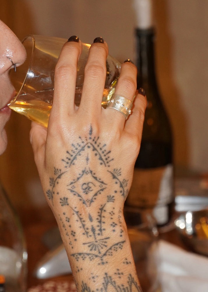 Person holding a glass of yellow liquid with a tattooed hand, wearing a silver ring