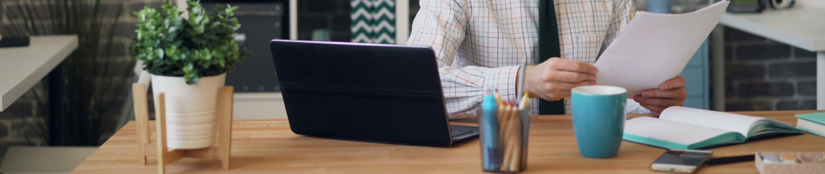 Man in shirt and tie working on desk, laptop, green plant, notepad and coffee can be seen