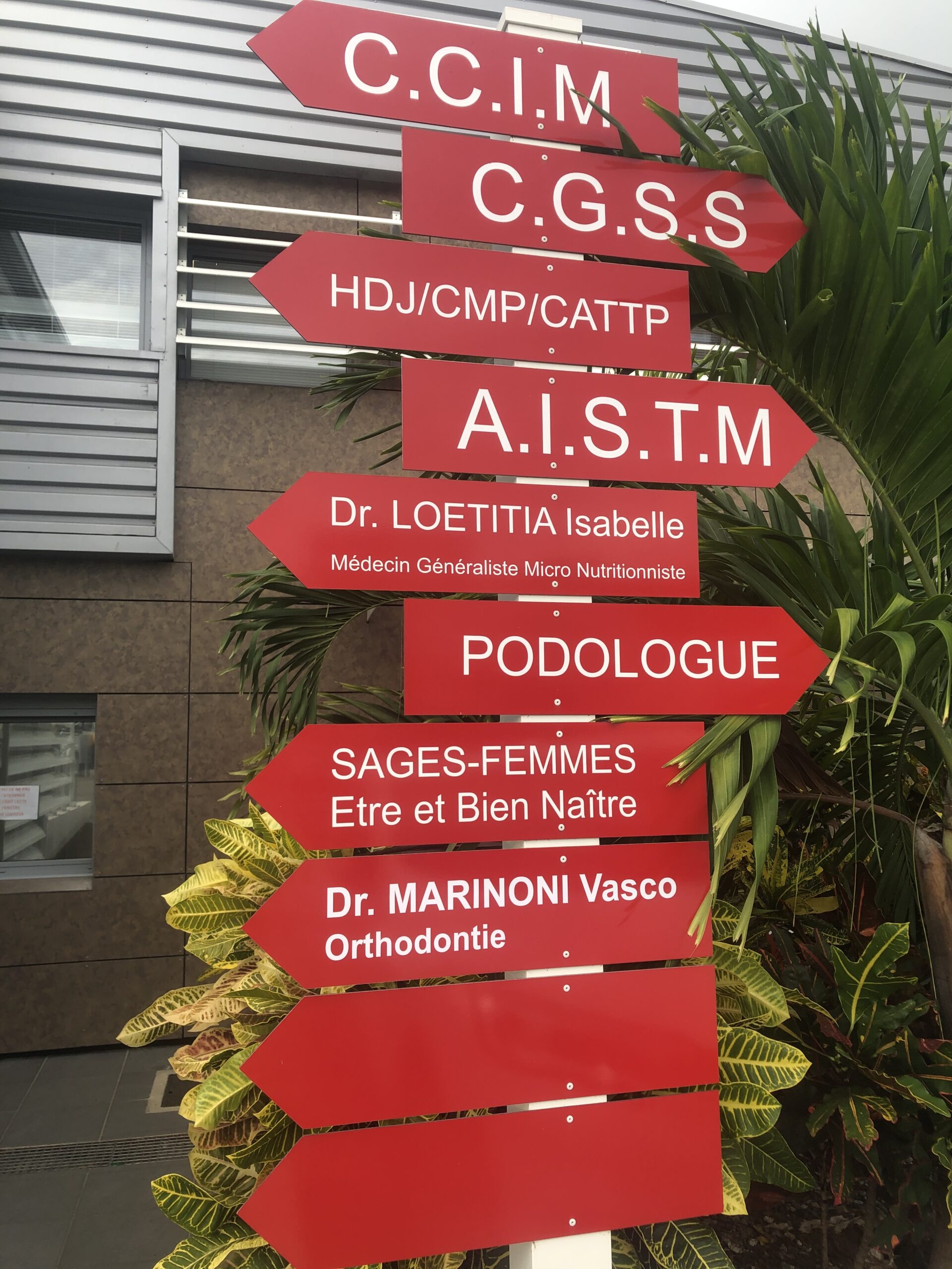 Des panneaux directionnels rouges avec du texte blanc indiquant les services médicaux, les médecins et les cliniques sont entourés de plantes vertes, devant la façade moderne d'un bâtiment.